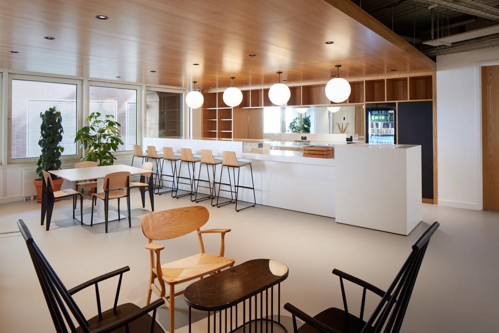 FT Architects - New Broadhouse - Project Image 5 Kitchen and bar area with a white counter, timber ceiling and globe pendant lights, with informal seating.