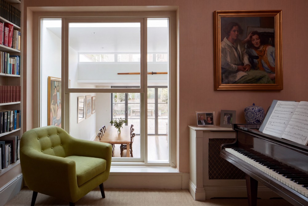 Notting Hill House — Music Room Dusky-pink music room with grand piano and green armchair, looking through an internal window to the garden extension